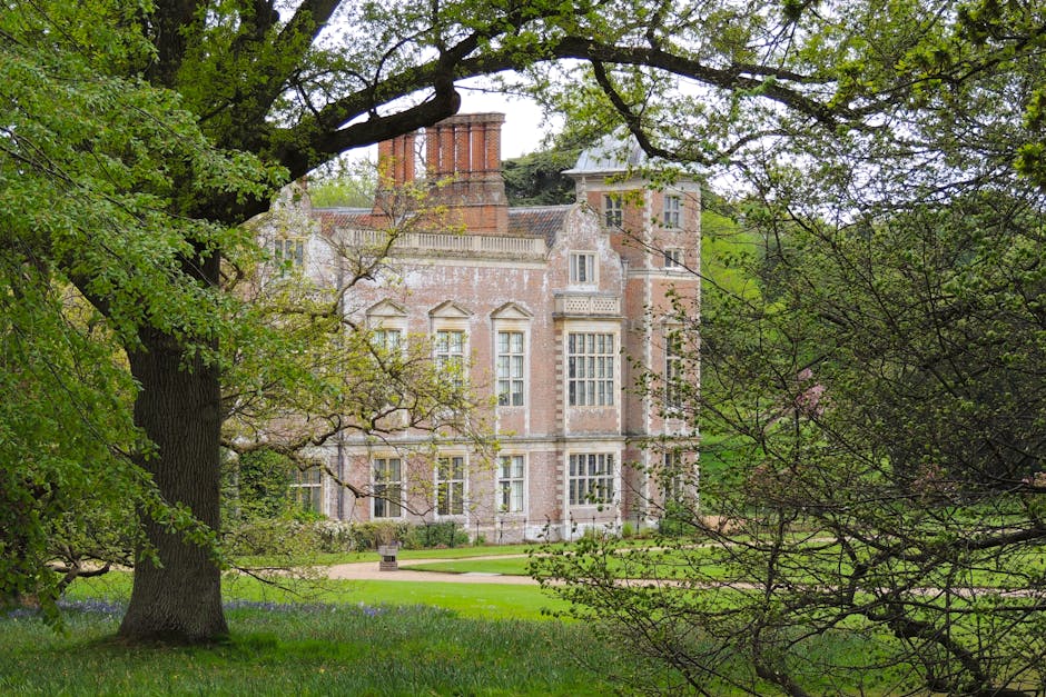 An aerial view of a expansive estate featuring a historic mansion surrounded by well-maintained lawn areas, winding pathways, and an abundance of mature trees and shrubs. The gardens display neatly tr